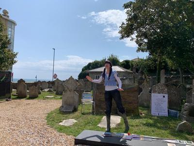 A person with brown hair in pigtails and a blue top and black trousers is frozen in place on grass in the middle on a sunny day in a graveyard 