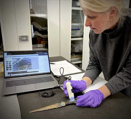 A person wearing purple gloves uses a small handheld tool to work on an dagger on a worktable while viewing a microscope image on a laptop in a lab setting.