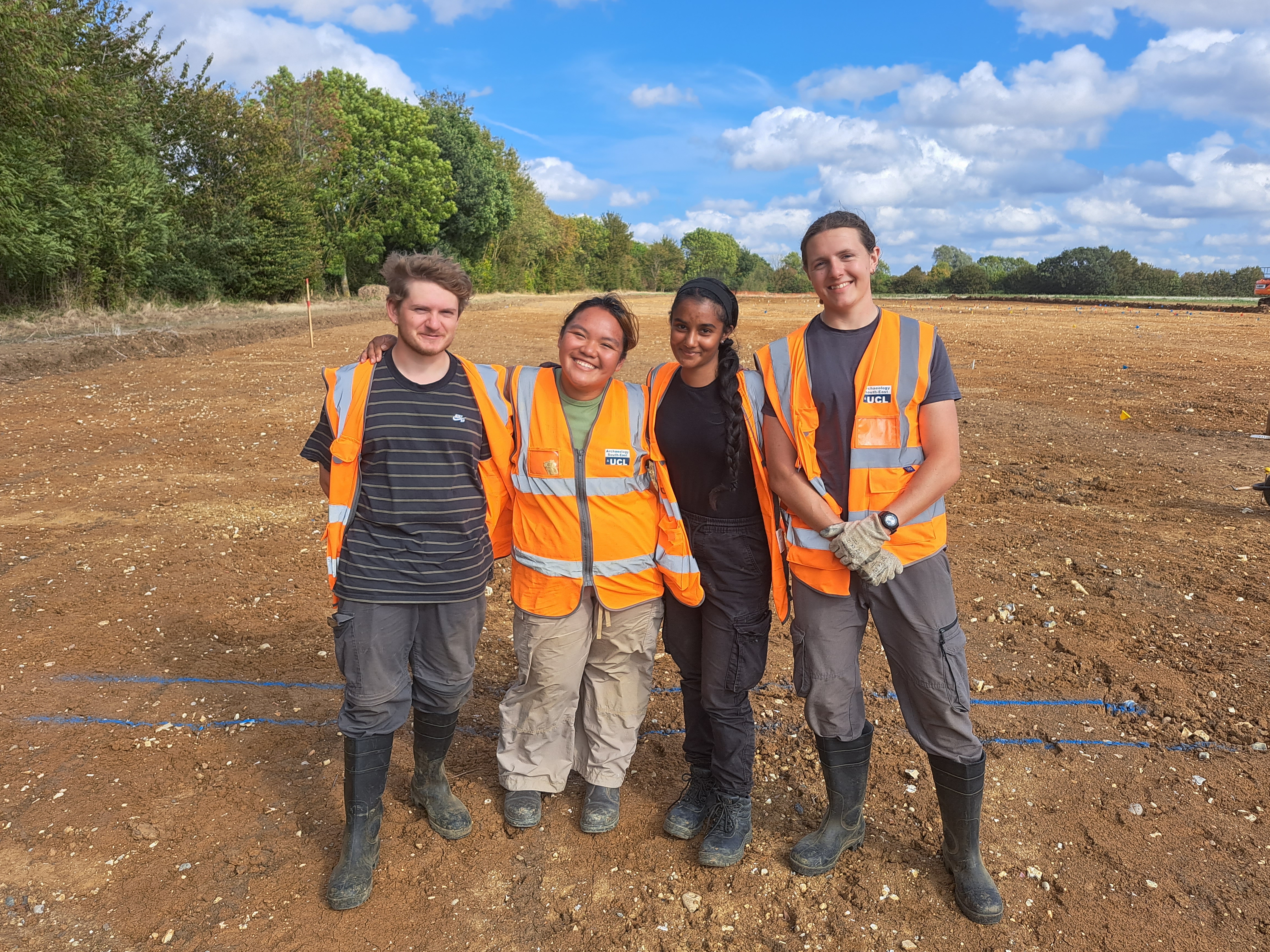 Four students pose together for a group photo on an archaeological site. They are all wearing high visibility orange vests.