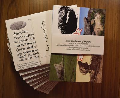 A stack of postcards fanned out on a dark wooden table, with one set on top that has a description of the 11 cards inside the set. 