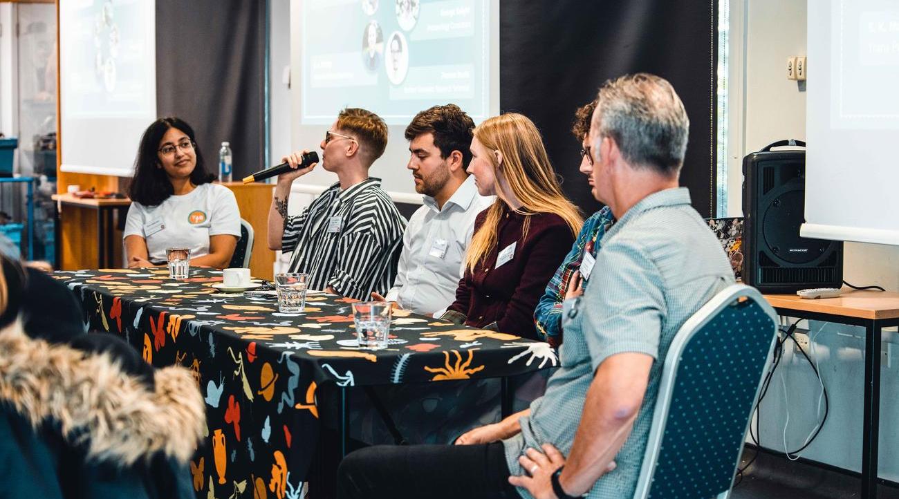 Six people sat in a row behind a table. One talking into the mic