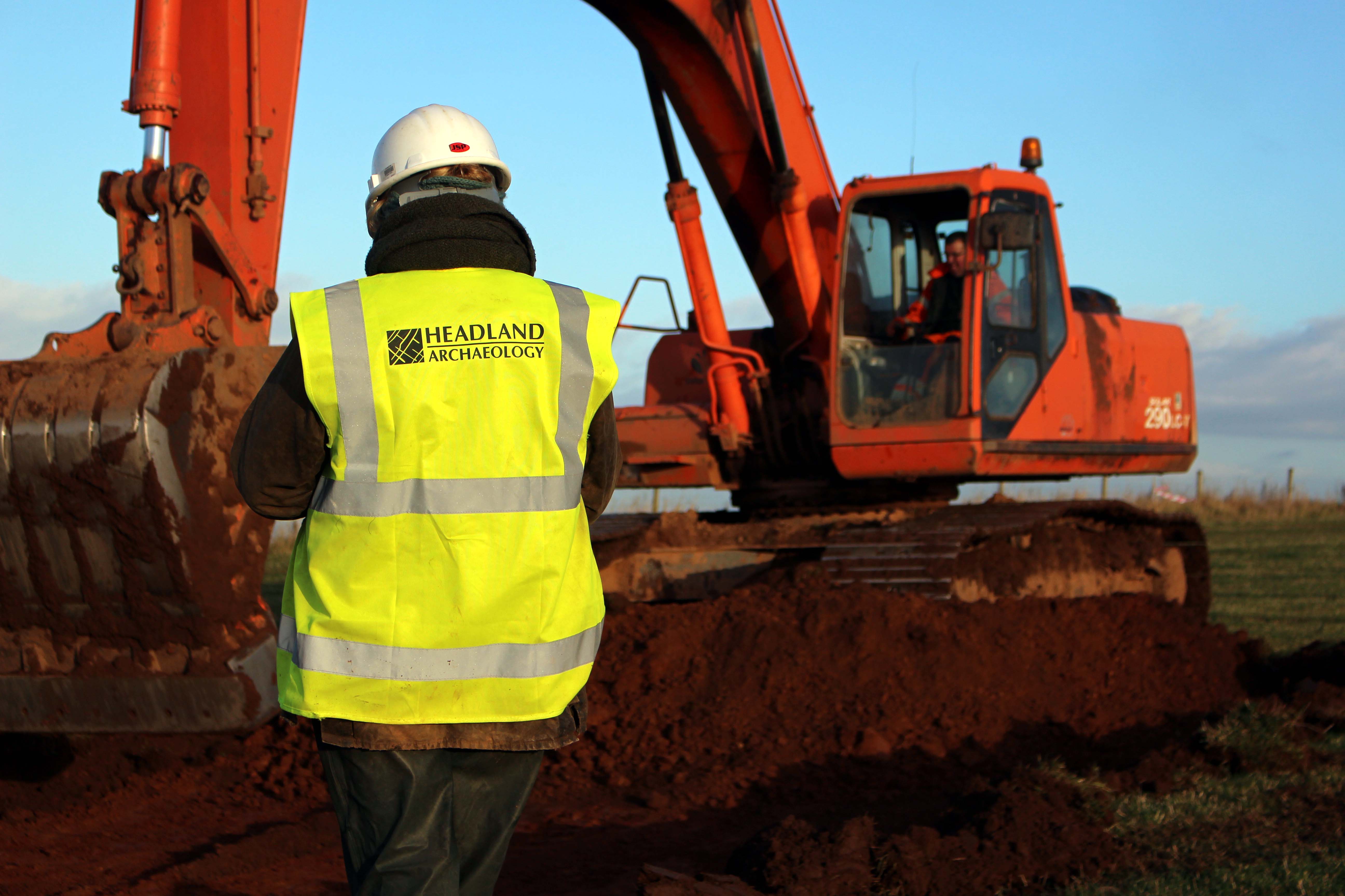 An archaeologist in a white hard hat and green hi vis jacket with Headland Archaeology logo on faces away from the camera.
