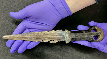 A gloved hand holds a small, corroded dagger with a ringed handle and worn blade, photographed on a gray surface.