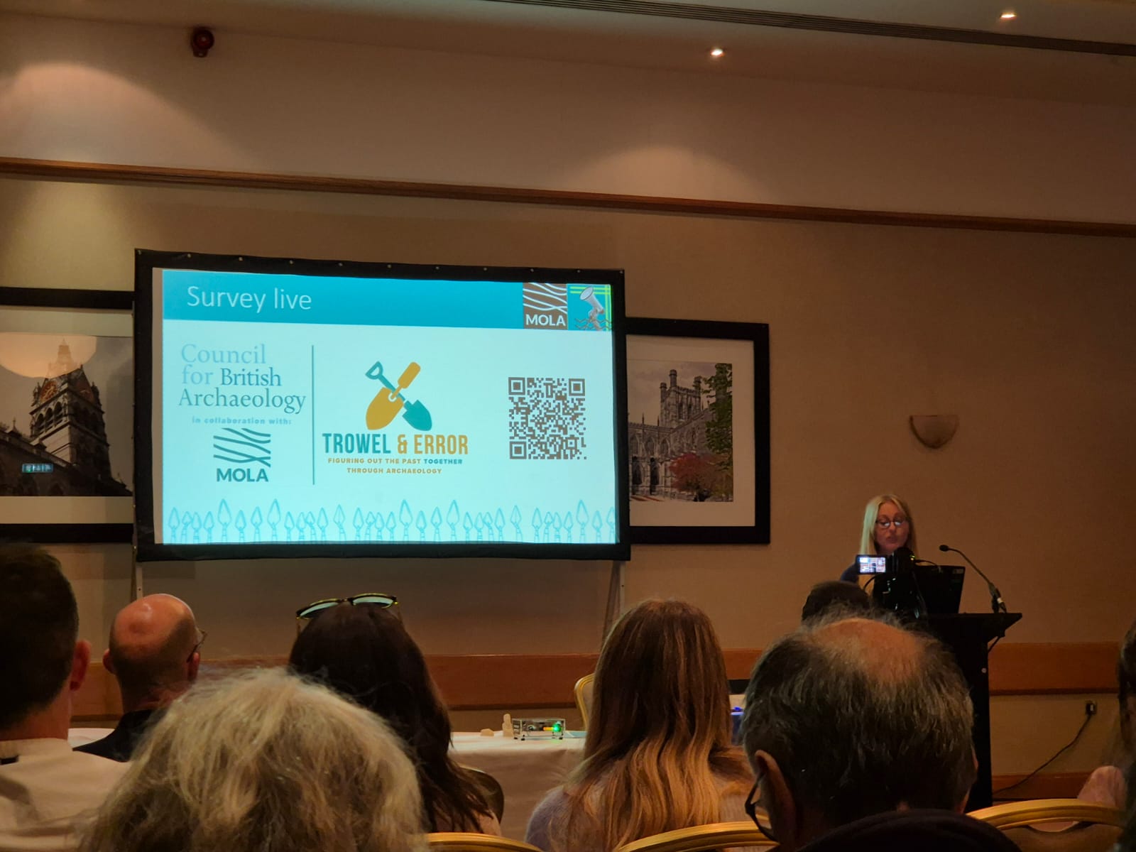 Young woman with blonde hair stood in front of a PowerPoint with the title 'Survey Live' and an audience in front of her.