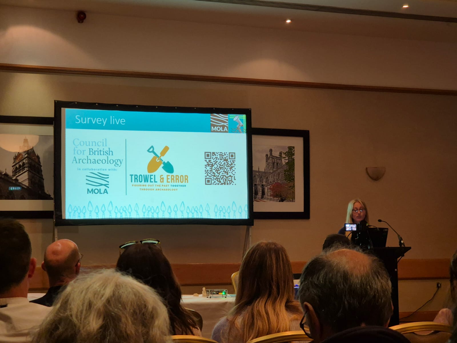 Young woman with blonde hair stood in front of a PowerPoint with the title 'Survey Live' and an audience in front of her. 