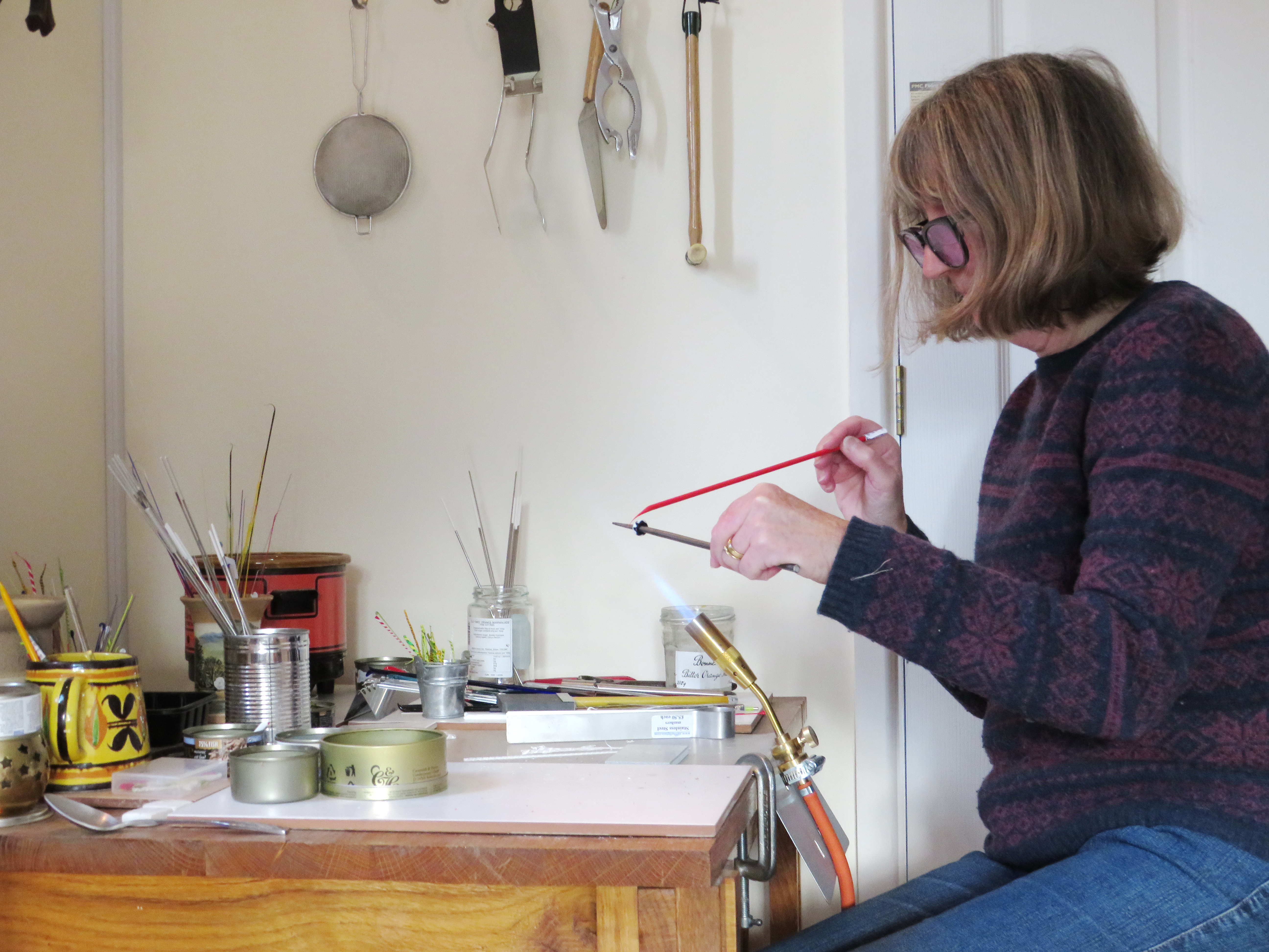  a person holding a flame tool and using a glass rod, they are sitting at a desk with lots of jars on