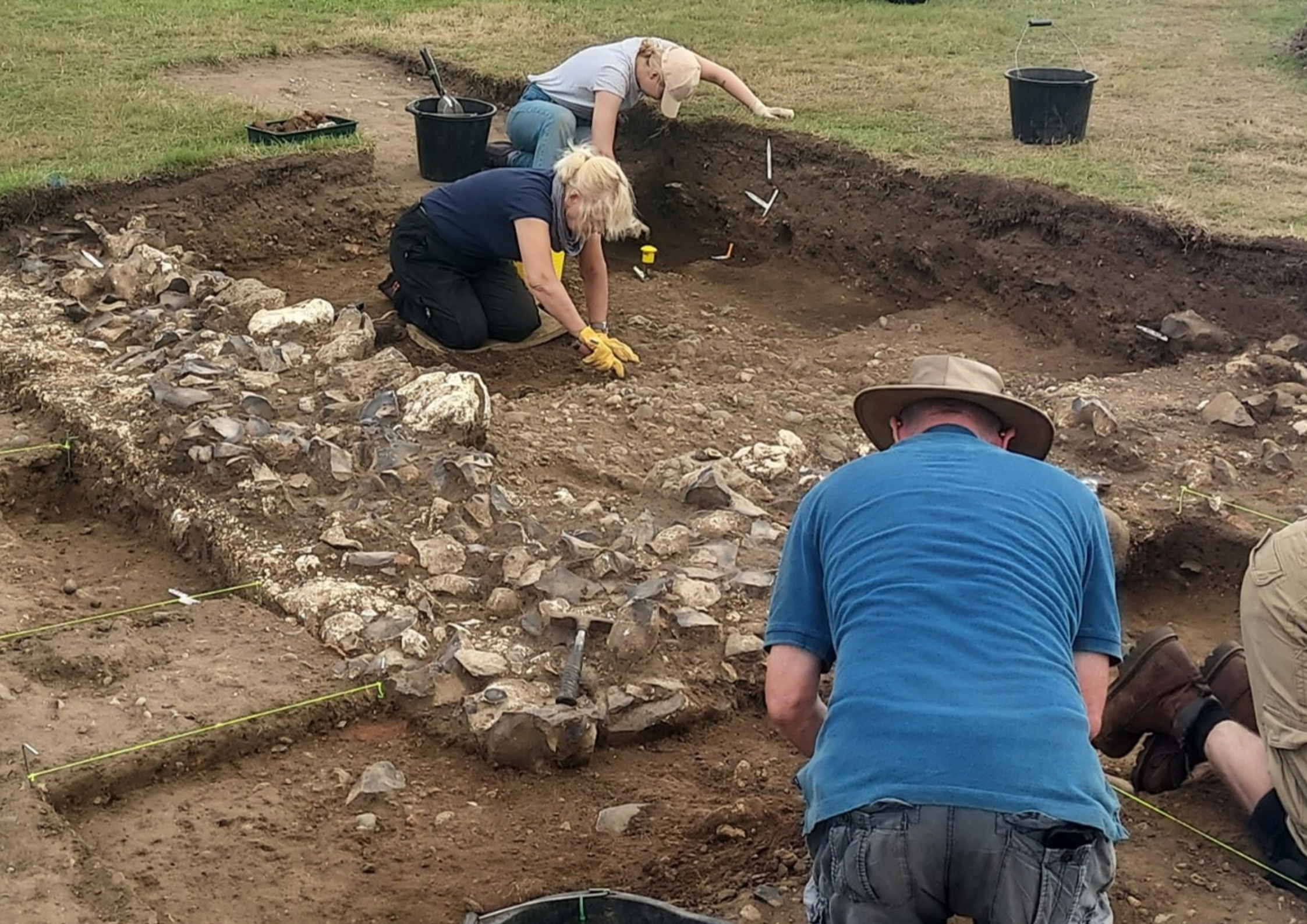 Three archaeologists kneeling down to unearth stone walls at an archaeological dig site. 