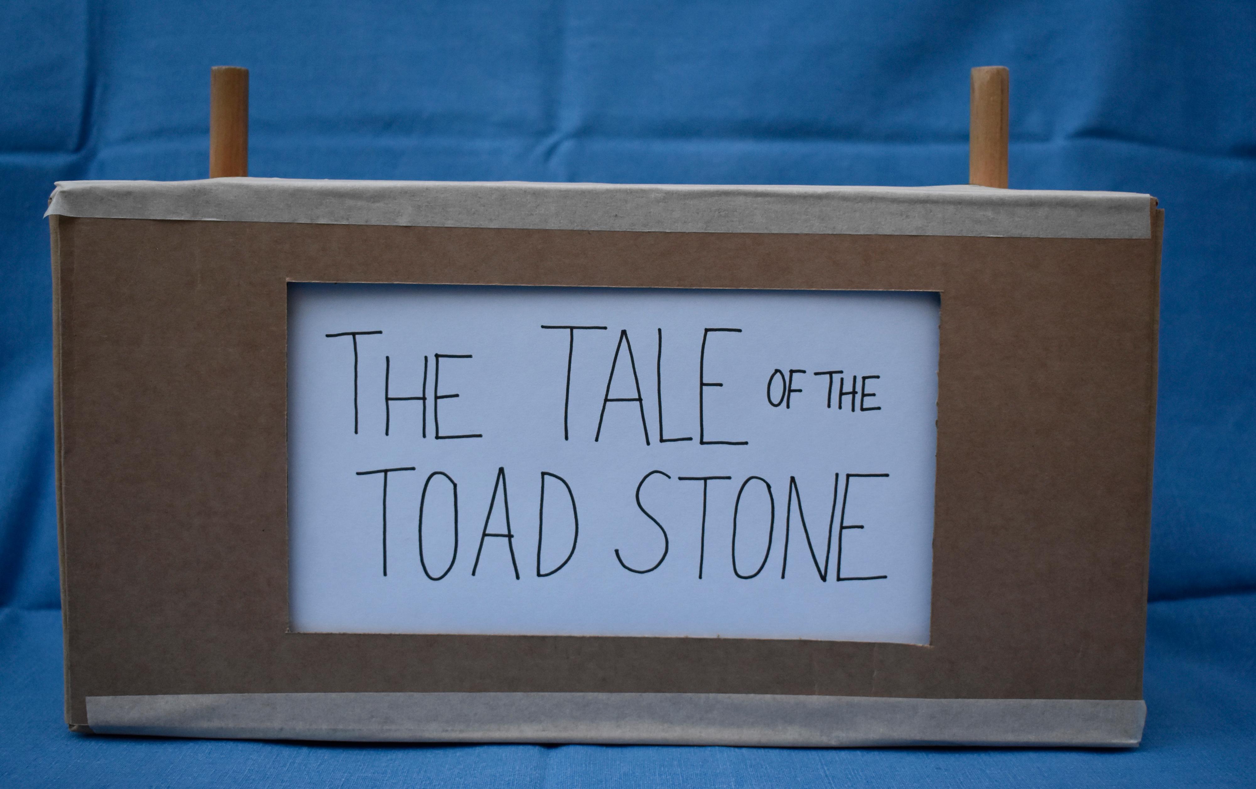 Hand made The Tale of the Toad Stone sign on a cardboard box with two wooden sticks sticking out the top with a blue sheet background